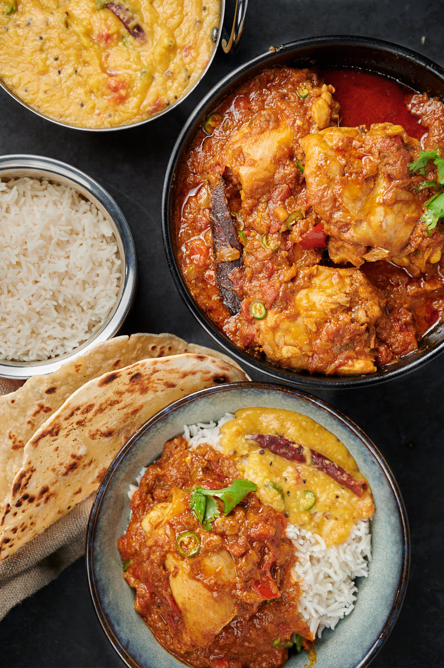 Simple chicken curry in a serving bowl with rice, paratha, and dal from above.