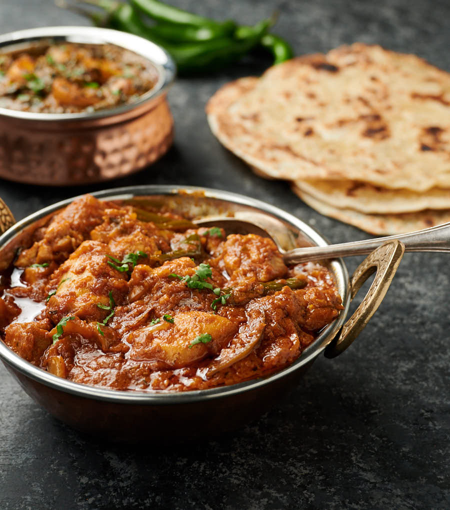 Chicken dopiaza curry in a bowl with serving spoon. Parathas in the background.