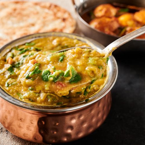 Spoonful of lentil curry with spinach with parathas and egg curry in the background.