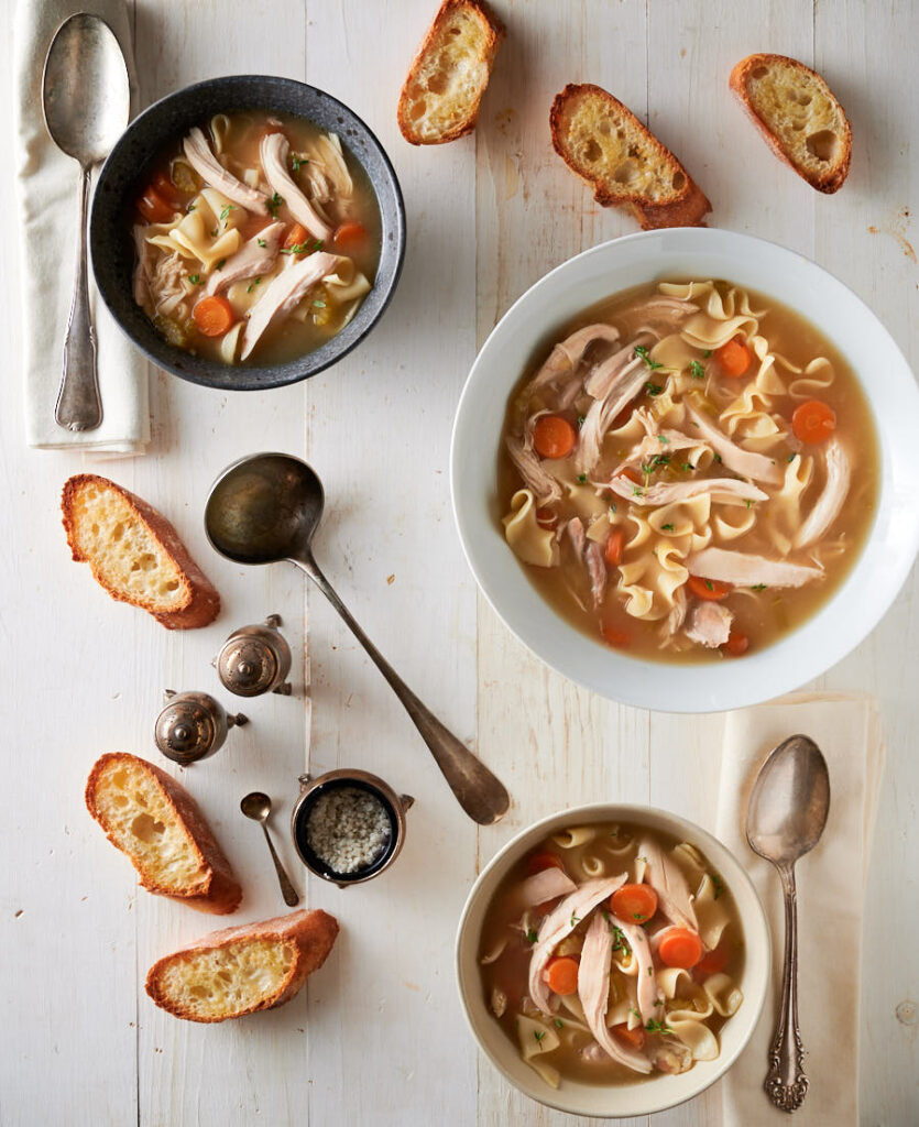 Table scene of homemade chicken noodle soup. Spoons, bread, napkins and ladle from above.