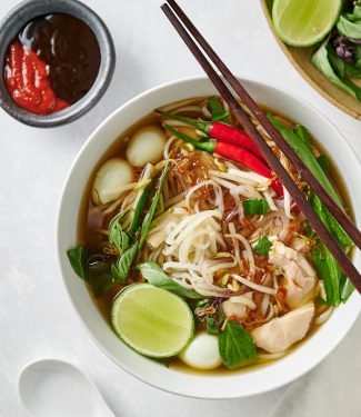 Bowl of pho ga with chopsticks with hoisin and sriracha side dishes.