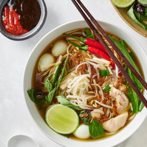 Bowl of pho ga with chopsticks with hoisin and sriracha side dishes.