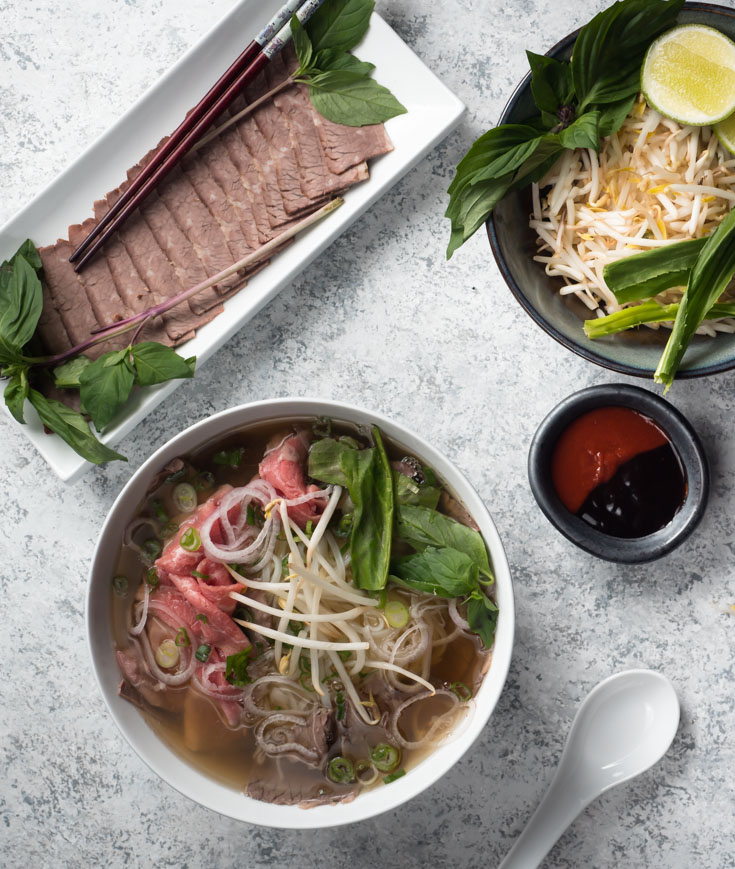 Bowl of beef pho, plate of brisket slices and garnishes from above.