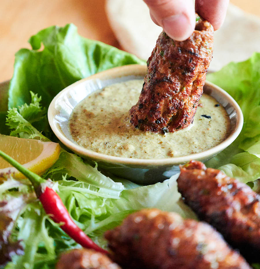 Seekh kebab being dipped into a bowl of yoghurt mint coriander dip.