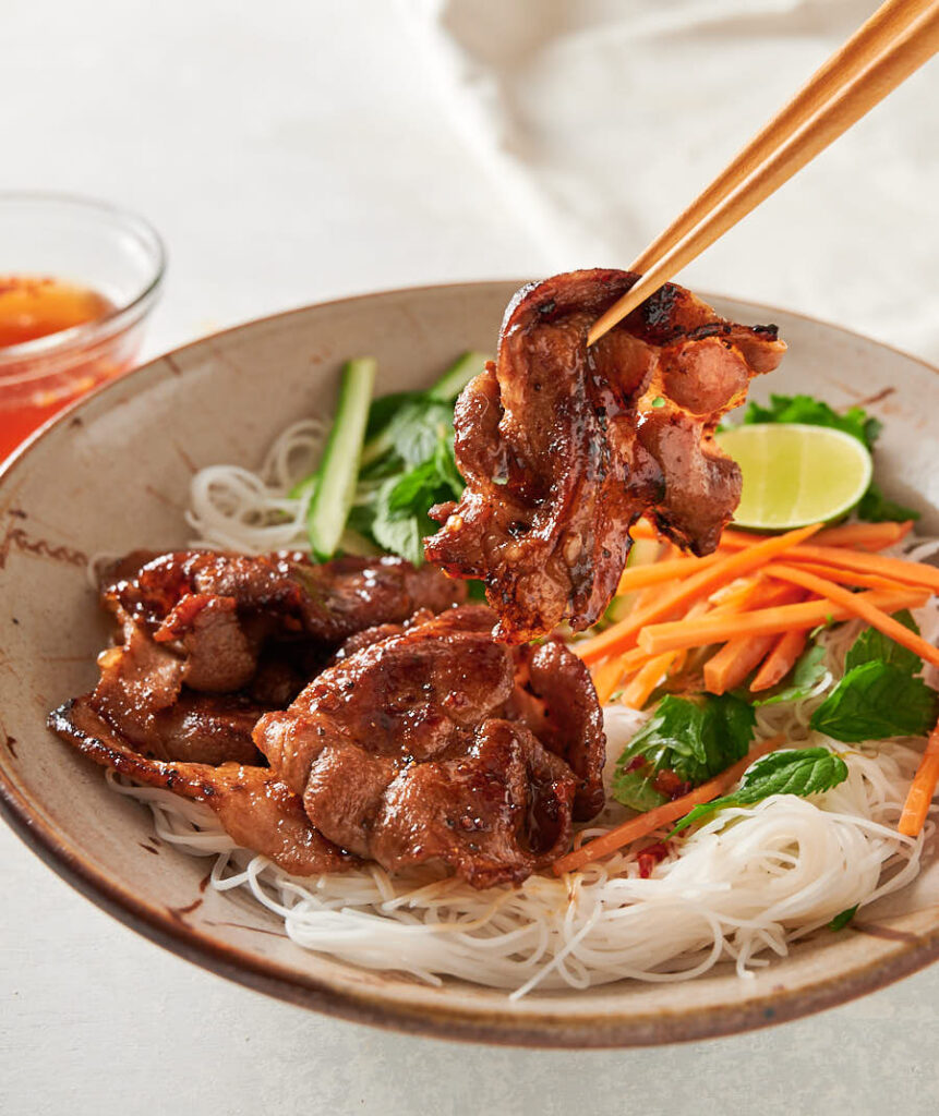 Chopsticks holding a piece of pork over a Vietnamese noodle salad bowl from the front.