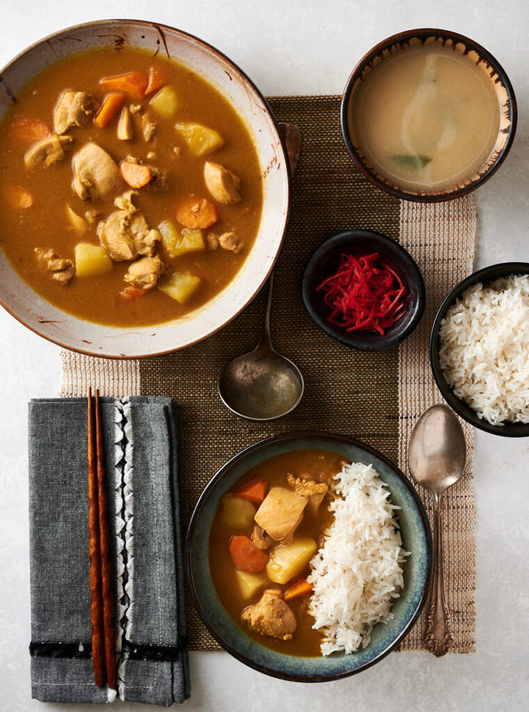 Table scene of Japanese chicken curry. Rice. Bowl of curry. Pickles. And plate with rice and curry.