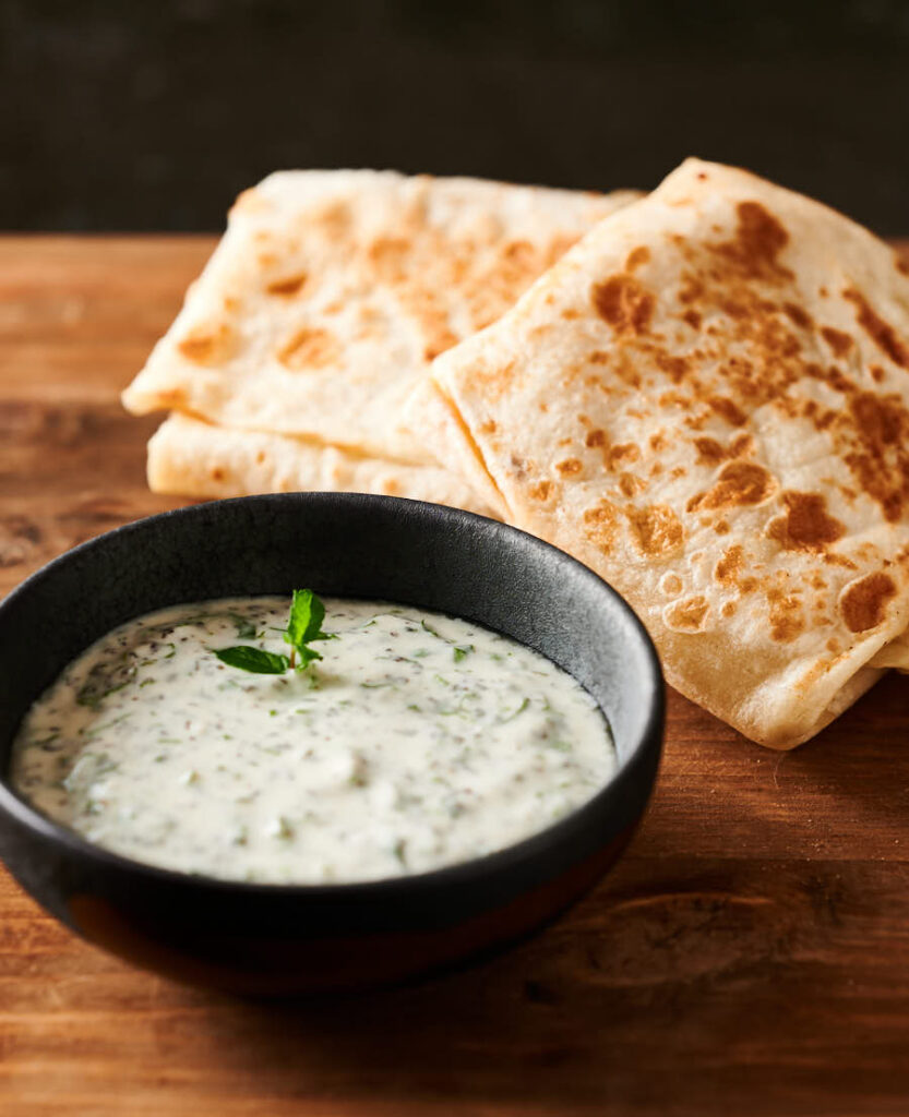 Bowl of mint raita in front of a couple mutton roti from the front.