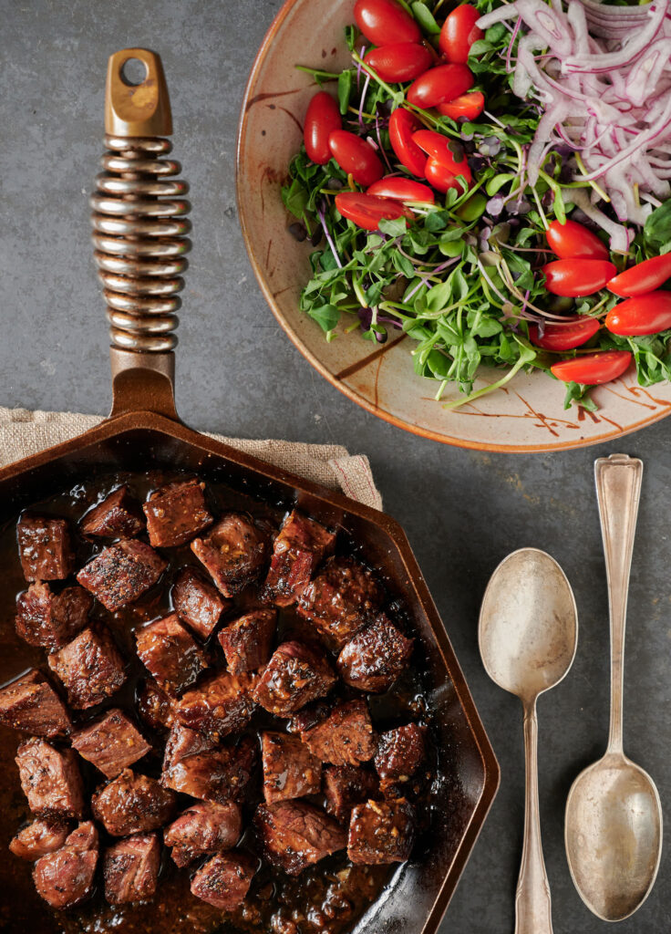 Cast iron pan of shaking beef next to a bowl filled with micro-greens, grape tomatoes and red onion from above.