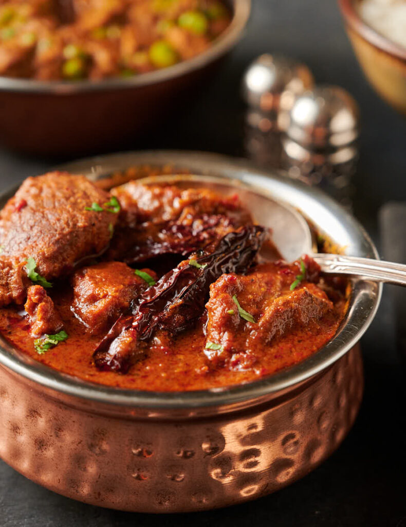 Close-up of an Indian copper serving bowl full of lamb vindaloo with a spoon.