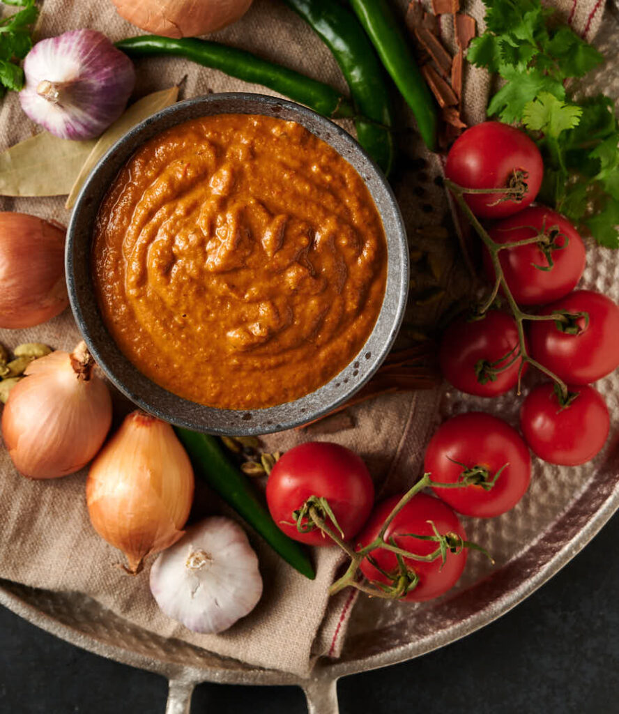 Overhead view of bold Indian hotel curry gravy surrounded by tomatoes, onions, garlic and green chilies.