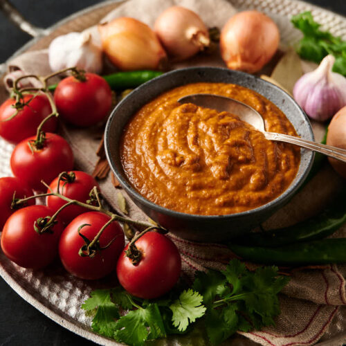 Indian restaurant curry gravy with a spoon sitting in it. Surrounded by tomatoes on the vine, onions, green chilies and spices. From the front.