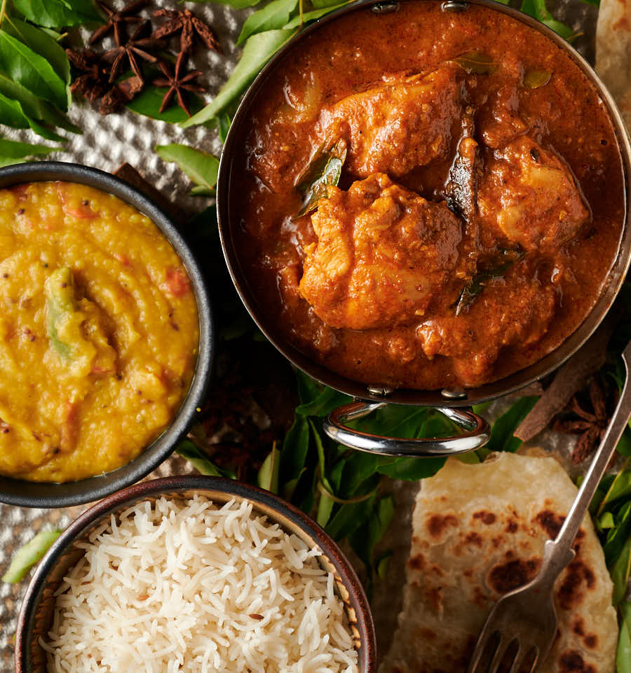 Closeup of chettinad chicken curry in a carbon steel bowl surrounded by fresh curry leaves.