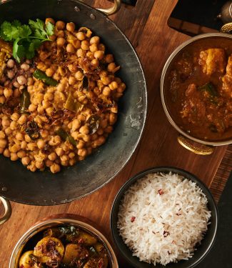 Table scene with serving bowls containing green chili chana achari, devilled potatoes, chicken dopiaza and rice from above.