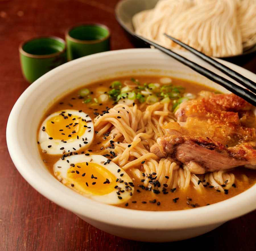 Bowl of curry chicken ramen, sake glasses and fresh ramen noodles table scene from the front.