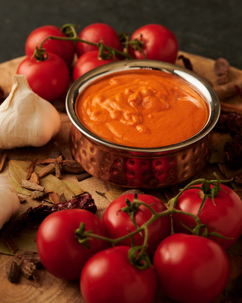 makhani gravy in a bowl surrounded by tomatoes, garlic and spices from the front.